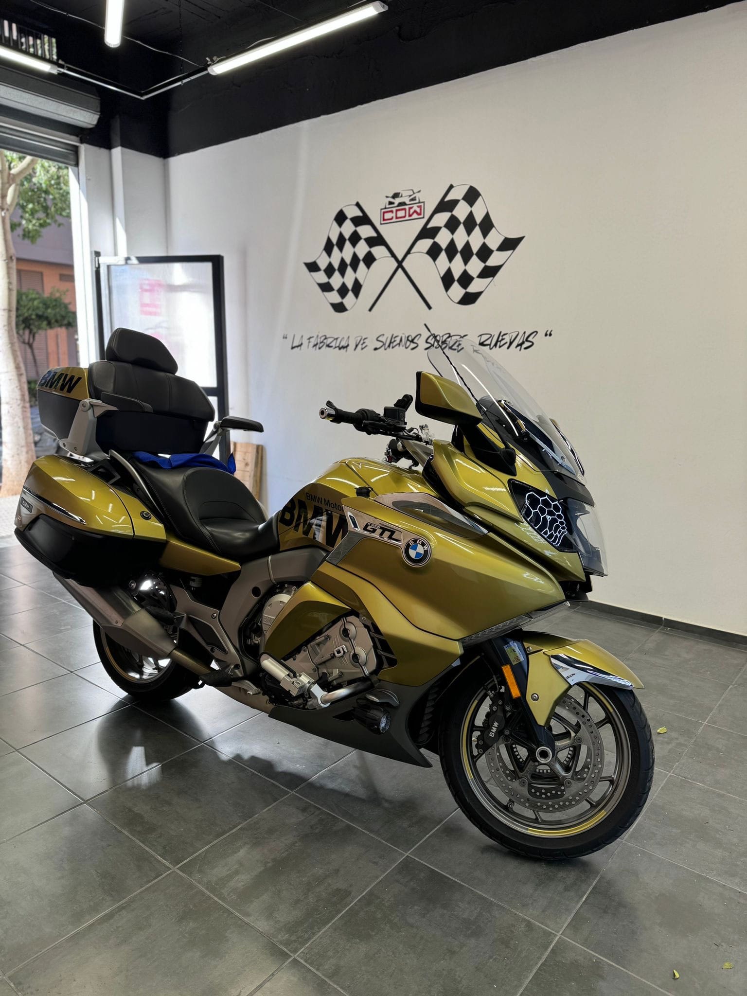 Motocicleta BMW K 1600 GTL personalizada con vinilo dorado metalizado dentro del taller CDW Llacer, con logotipo de banderas a cuadros en la pared.
